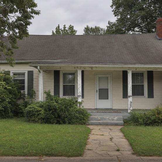 Older single-story house with worn porch trim, overgrown shrubs, and a cracked front walkway